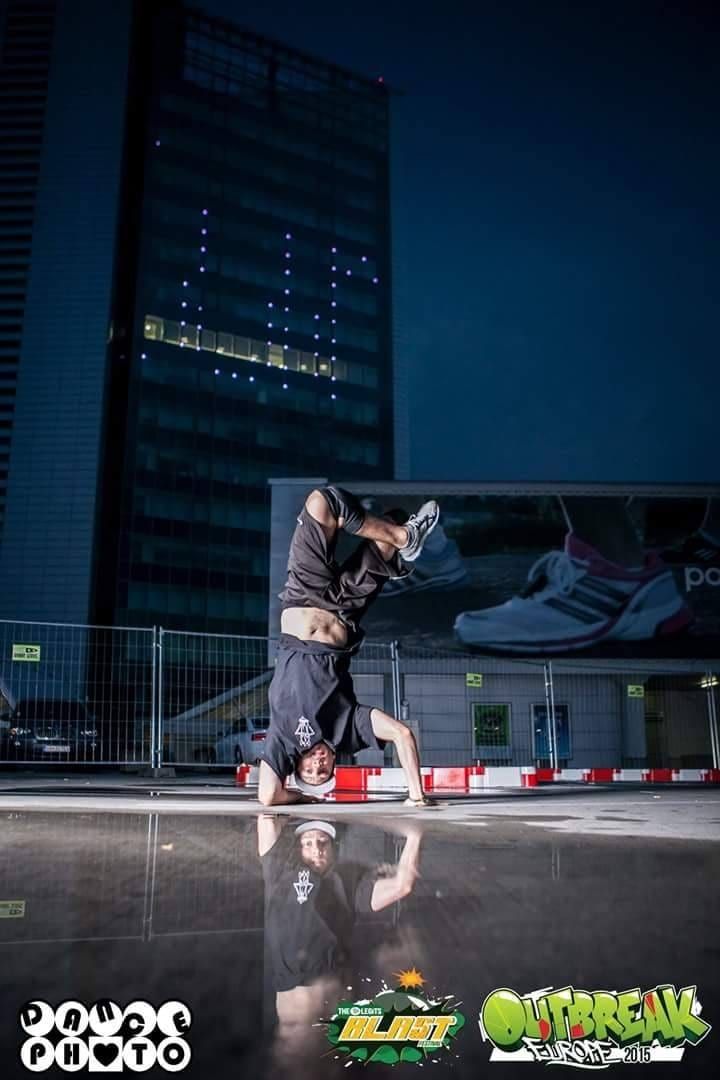 A dancer holding an inverted freeze on wet pavement at dusk, a tower block lit behind him
