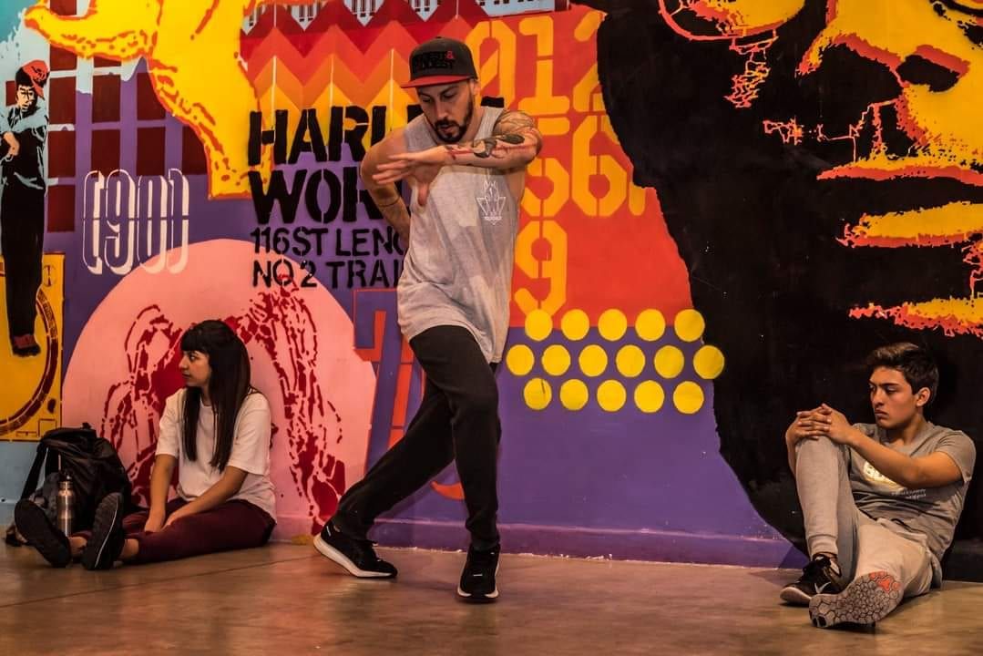 A dancer mid-move in front of a large graffiti mural while other dancers sit watching along the wall