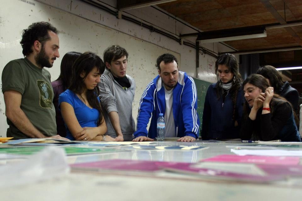 Students gathered around a studio table covered in large printed layouts, discussing the work