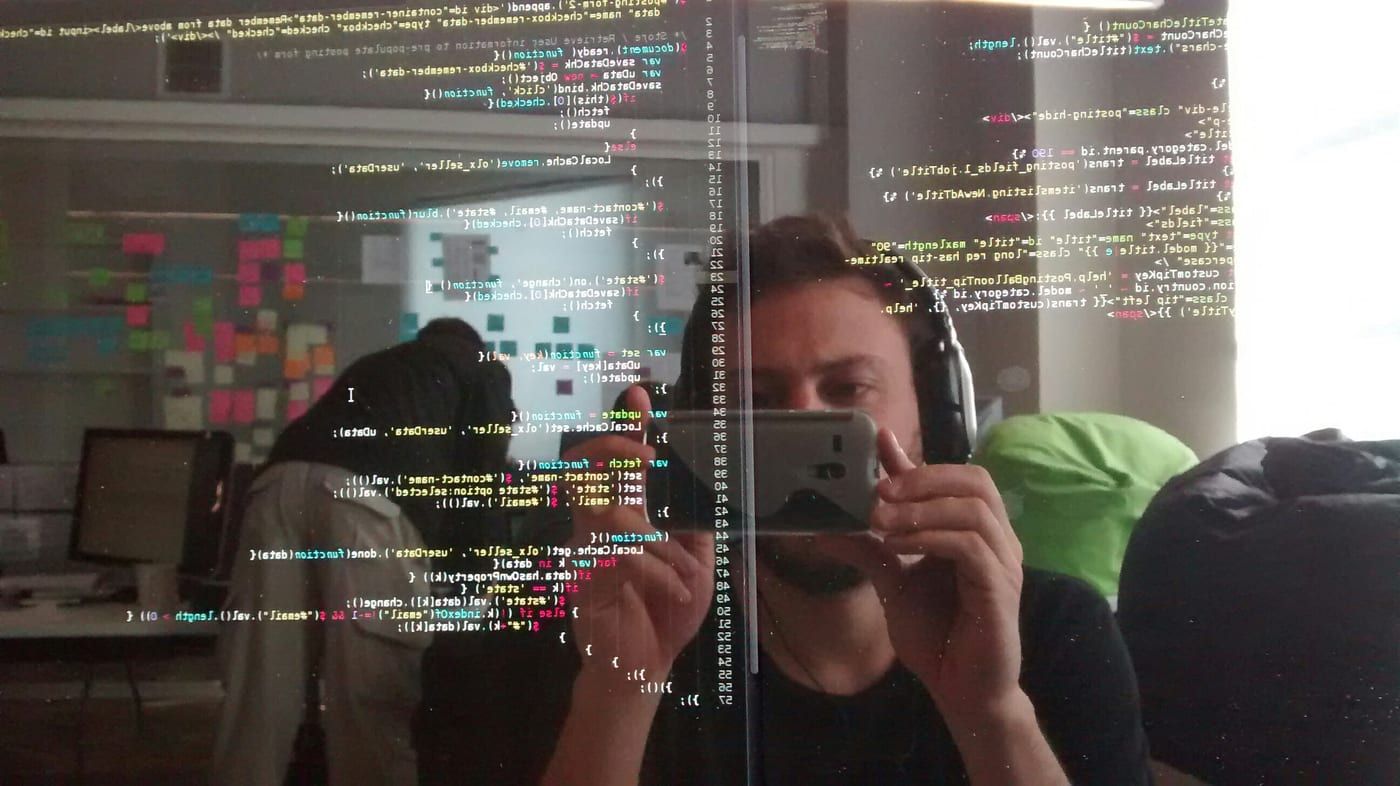 A developer at a desk seen reflected in a monitor filled with code, a wall of sticky notes behind him