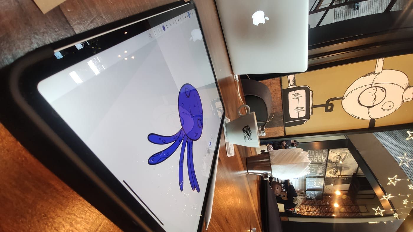An iPad propped on a desk showing a finished blue character illustration, a café interior behind it