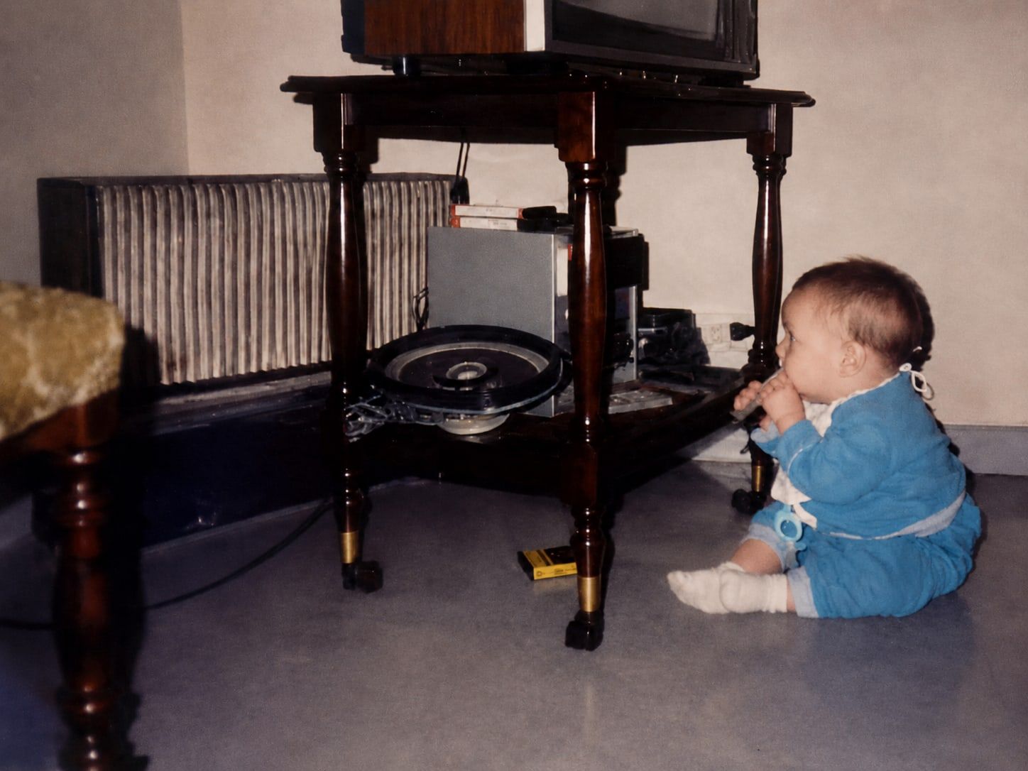 A baby sitting on the floor facing a wooden stand, a record player and a tape deck on its lower shelf and a television on top