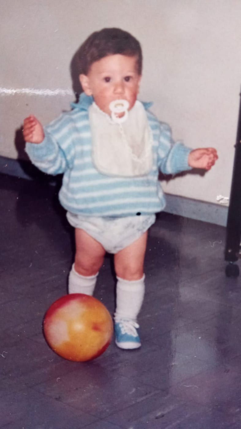 A toddler in a striped jumper standing over an orange ball on a tiled floor