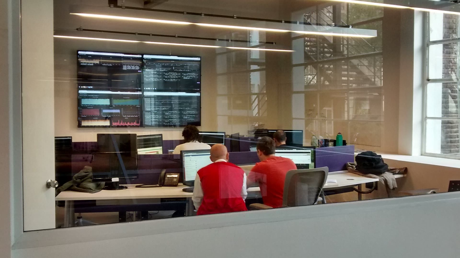 OLX monitoring room seen through a glass wall, with staff working below a four-screen systems dashboard.