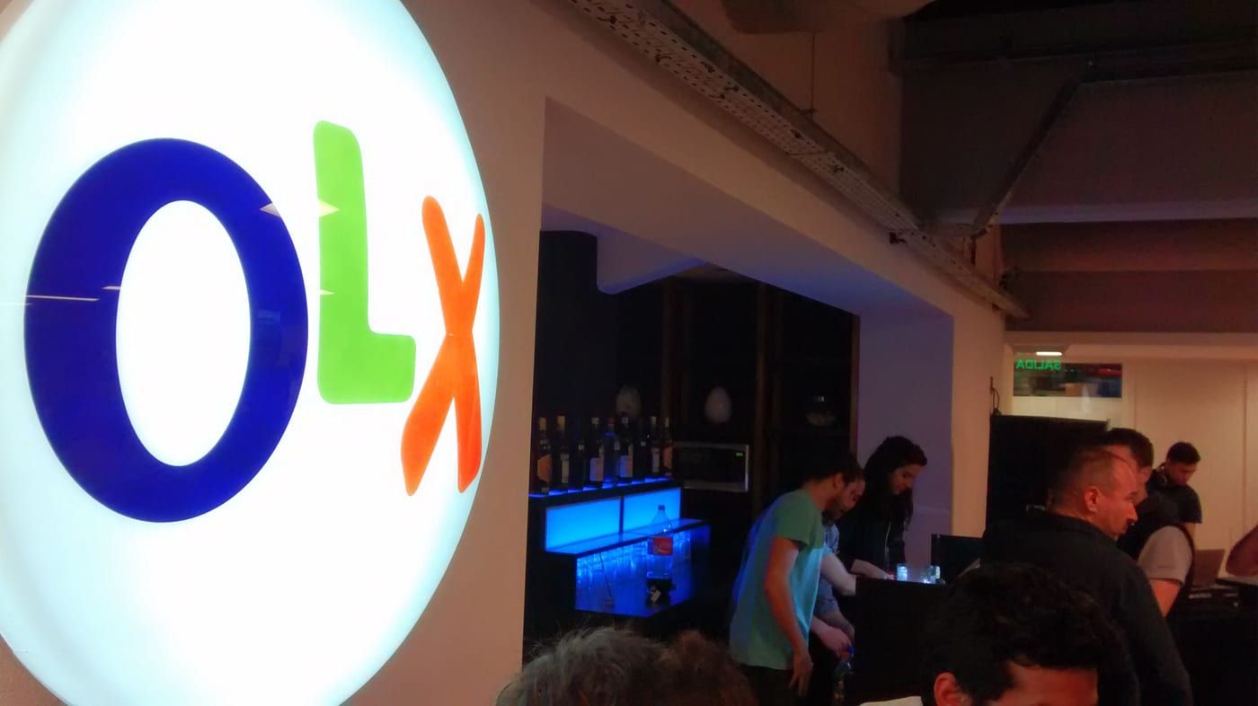 An illuminated OLX logo mounted in an open-plan office with people working at desks behind it