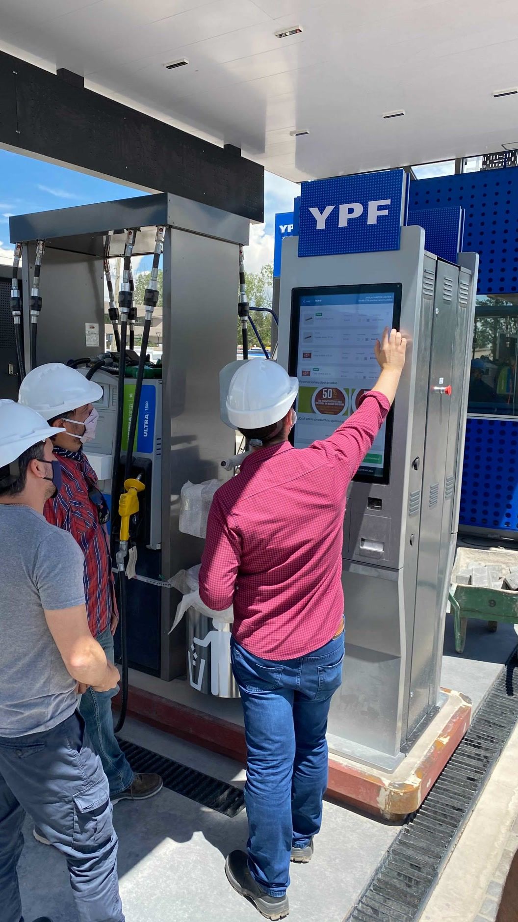 Four people in hard hats testing the multi-item catalog on a kiosk installed beside a fuel pump.